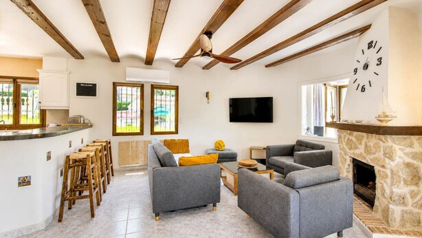 A living room with grey sofas, a TV, a stone fireplace, and wooden beams on the ceiling.