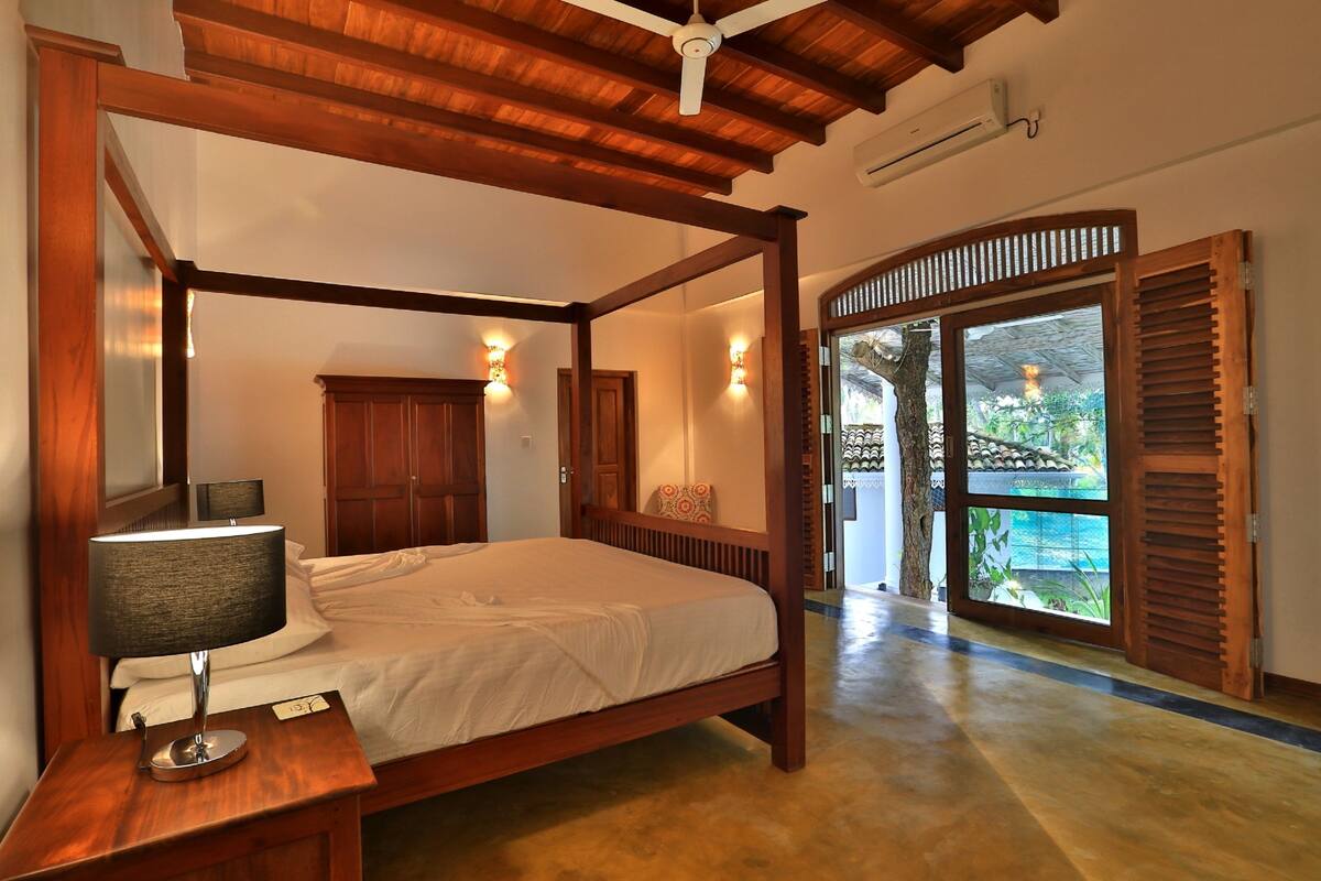 Uma cama de dossel com uma mesa de cabeceira, um abajur e vista para o exterior através de uma porta de vidro.