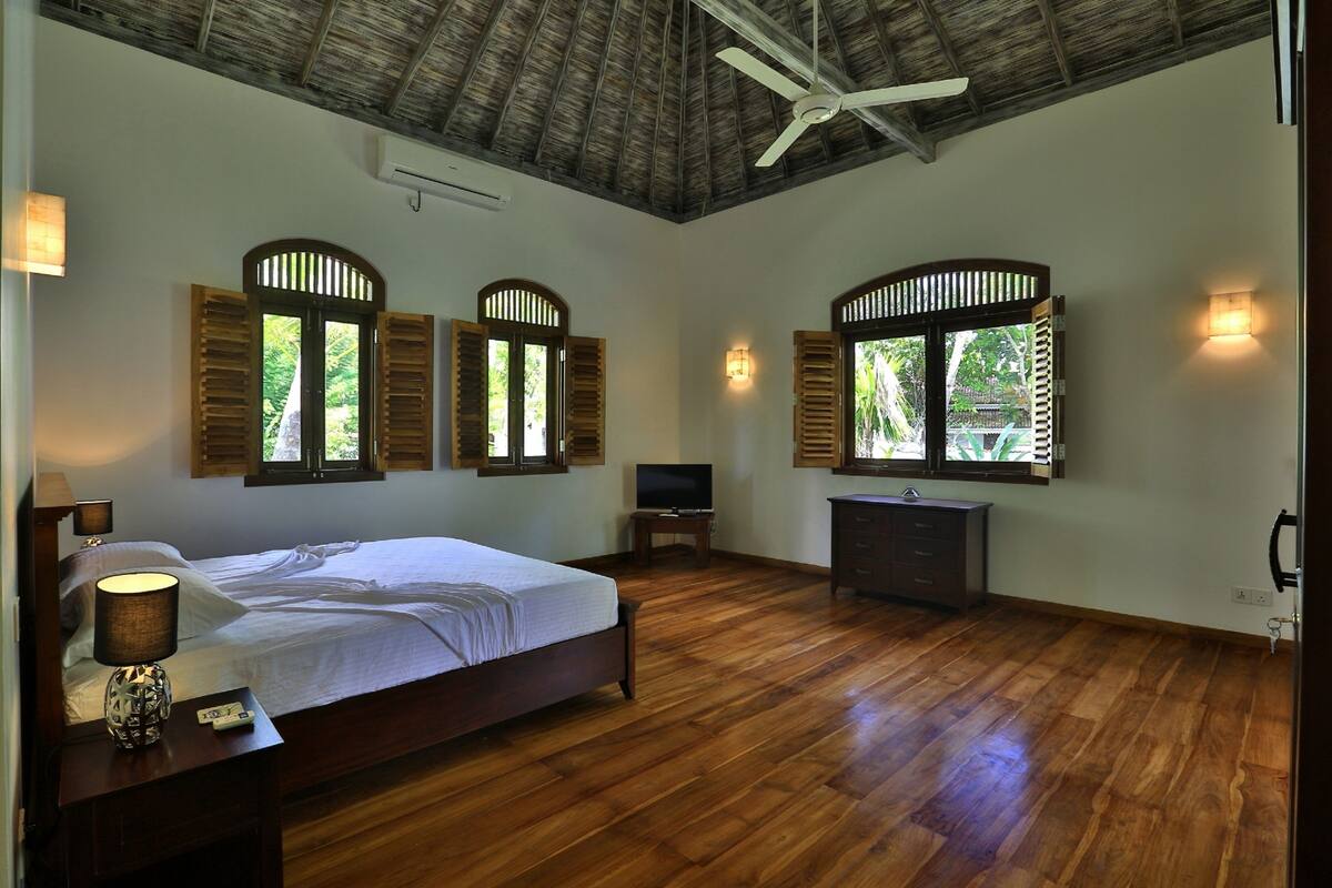 Quarto com piso de madeira, cama, televisão e duas janelas com persianas.