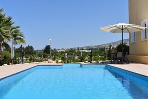 Une piscine rectangulaire à l’eau d’un bleu limpide, entourée d’un petit muret en pierre et de palmiers. On y trouve également un espace patio avec des meubles d’extérieur et un grand parasol.