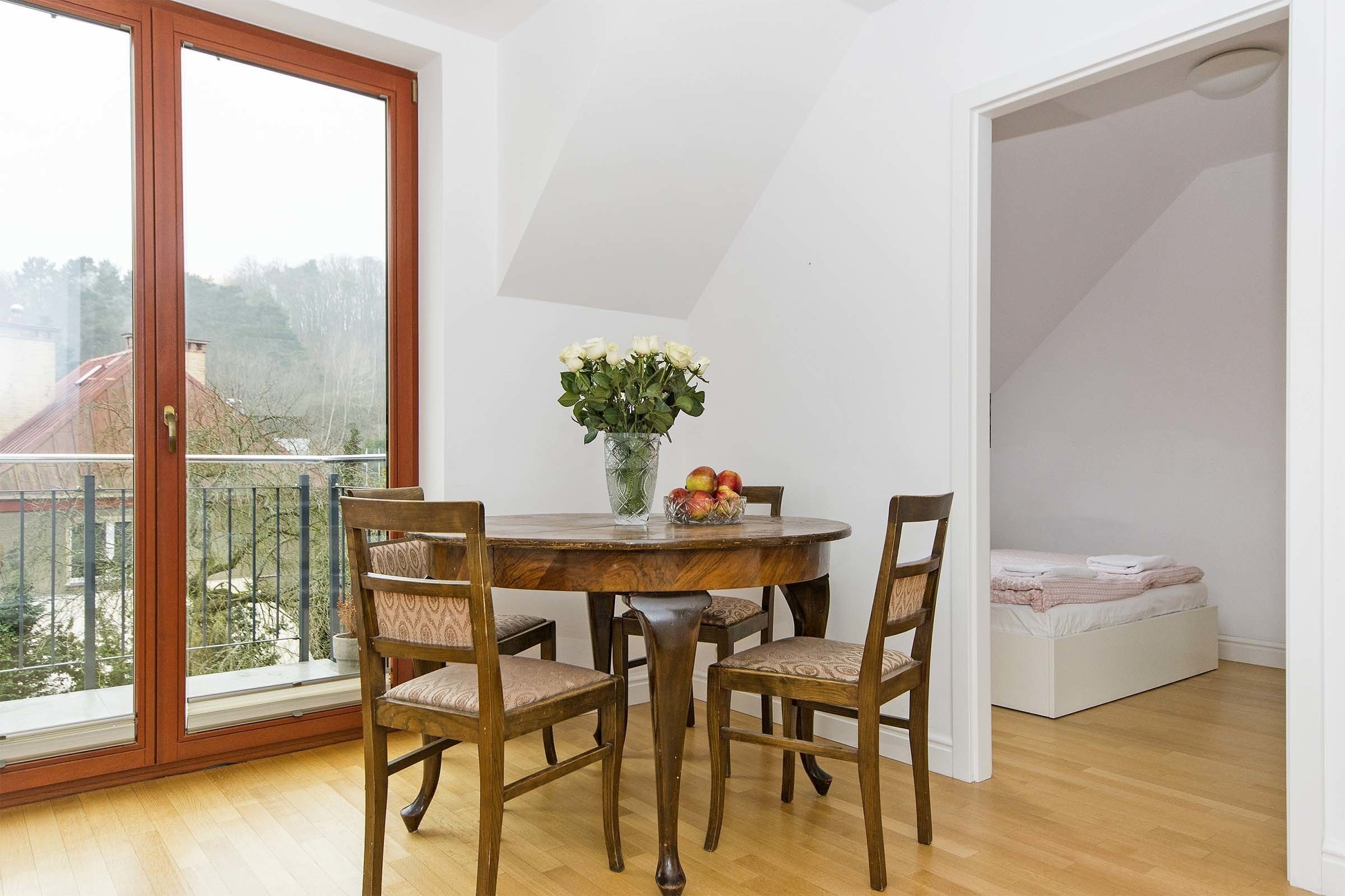 Un'area pranzo con un tavolo rotondo in legno, quattro sedie, un vaso con fiori e una ciotola di frutta. Si vede un balcone con ringhiera attraverso la porta vetrata.