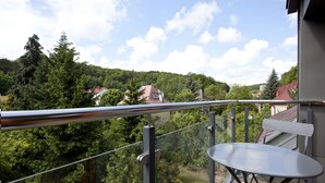 Un balcon doté d’une balustrade en métal, d’une petite table et offrant une vue sur une colline boisée parsemée de maisons.