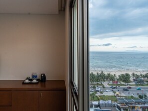 A room with a view of a beach and ocean, a wooden desk with a tray of items, and a window with a view of a busy street. - Tamarind Hotel Da Nang (Da Nang)