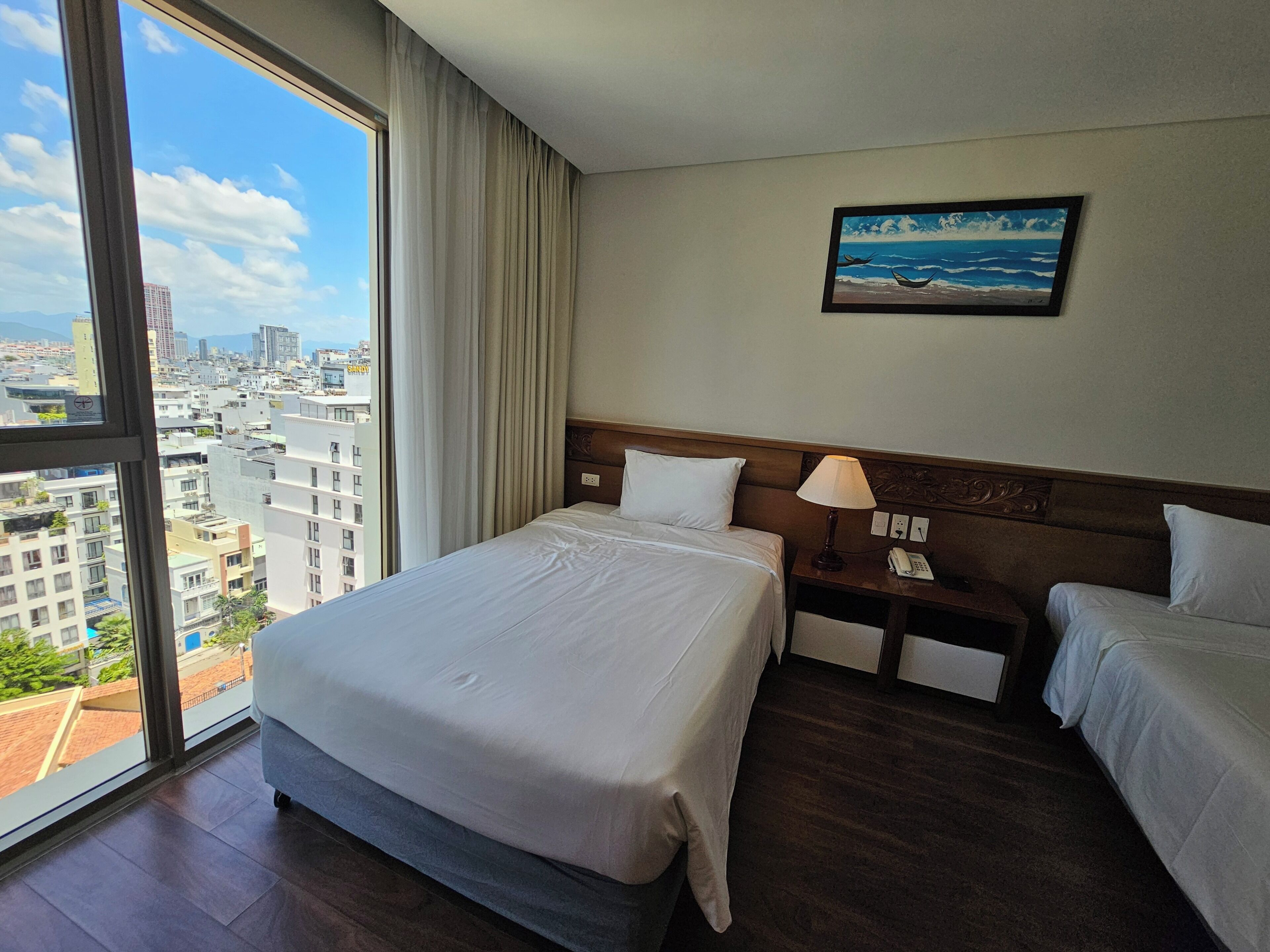 A hotel room with a large bed, a bedside table with a lamp, a phone, and a painting of a beach scene.