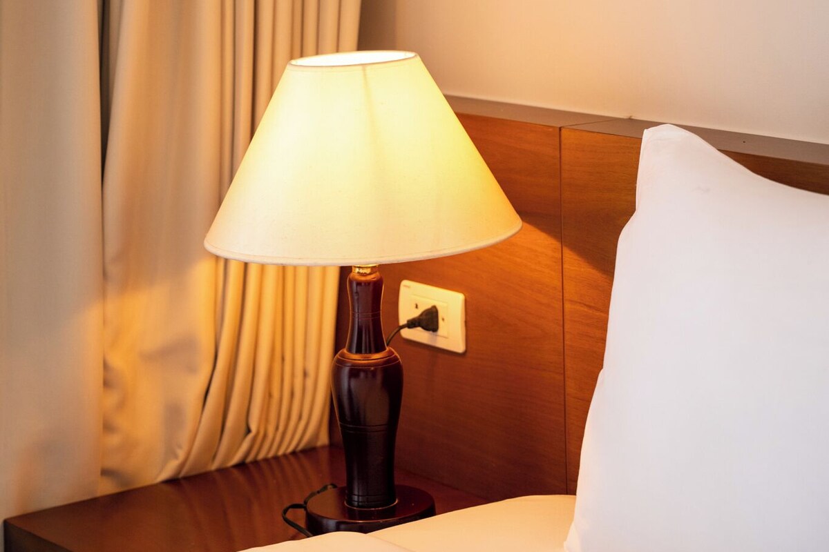 Habitación de hotel con una cama, una lámpara, una mesita de noche de madera, un almohadón blanco y una cortina.