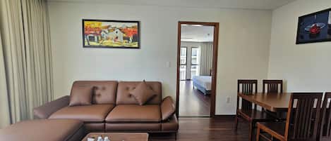 A modern hotel room with a brown leather sofa, a wooden coffee table with water bottles, a dining table with chairs, and a painting on the wall.