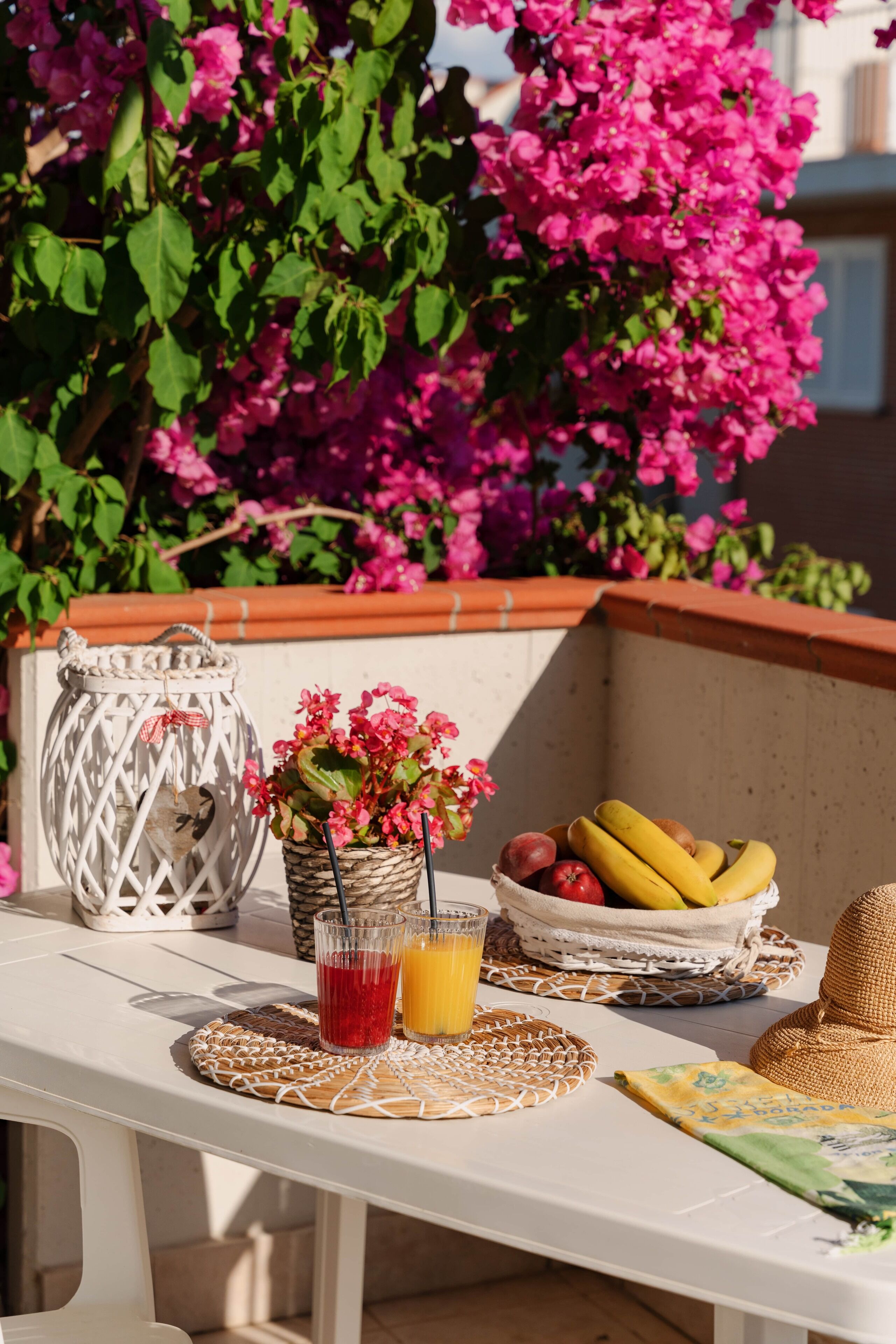 A table with a basket of fruits, a woven mat, a glass of red drink, and a wicker basket with flowers.