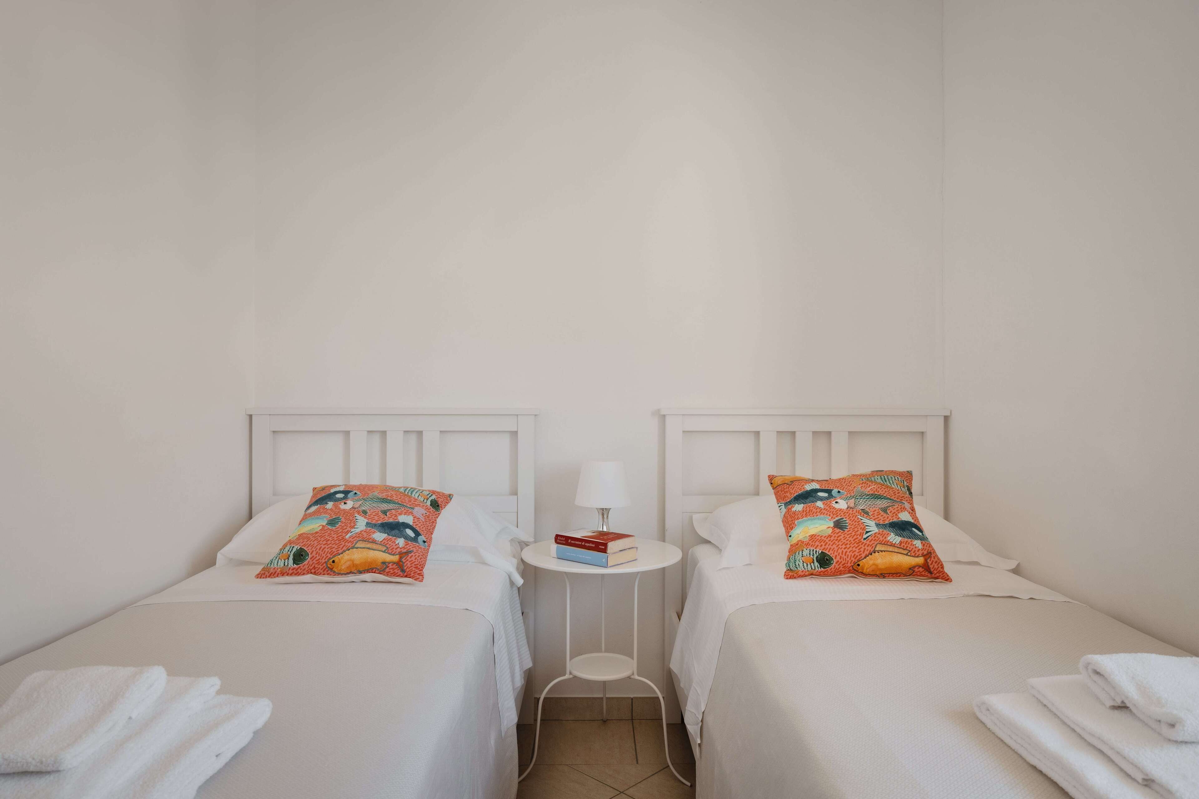 Una habitación de hotel con dos camas individuales, cada una con cabecero blanco, una mesita de noche pequeña con lámpara y un cojín colorido.