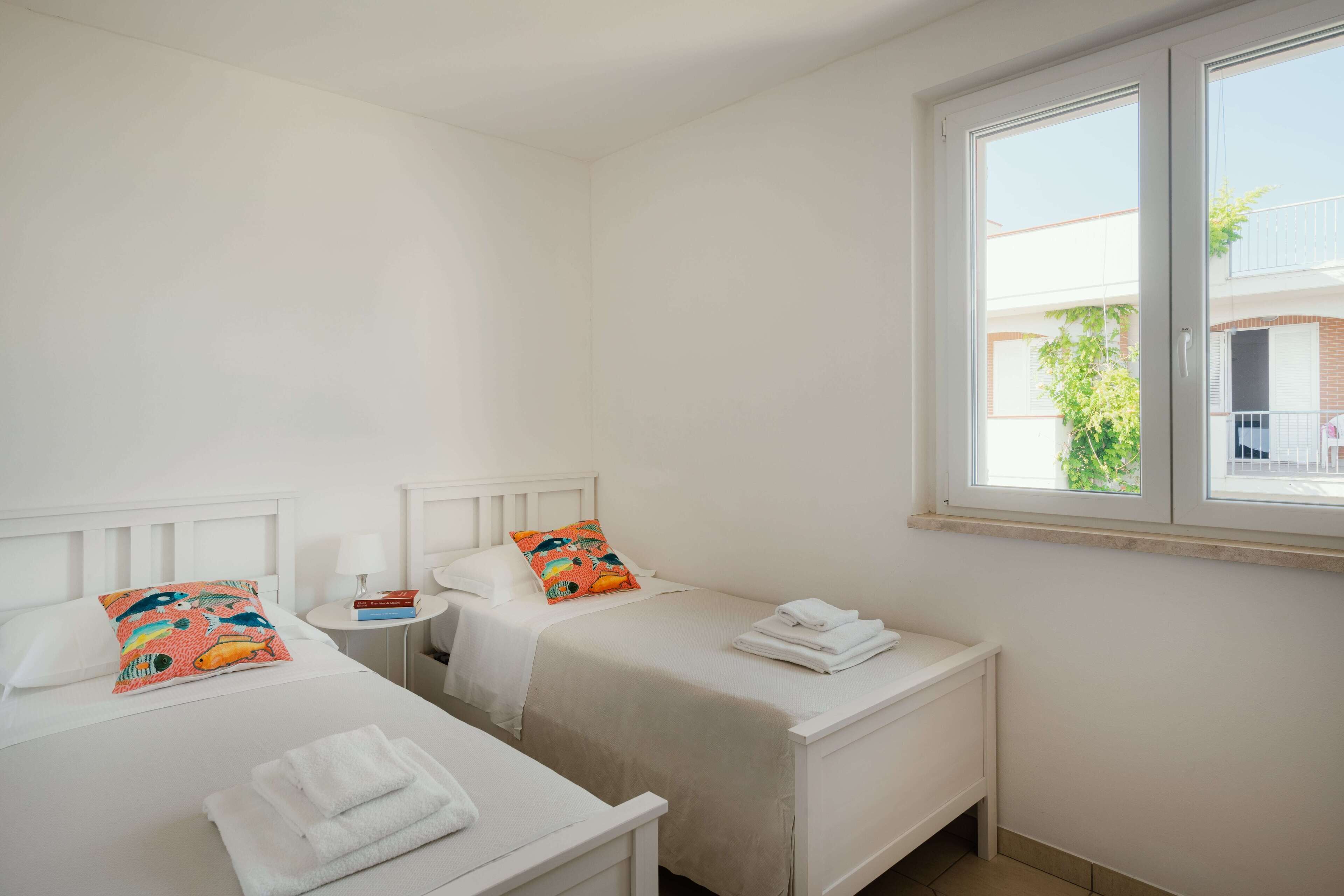 Habitación de hotel con dos camas individuales, una mesita de noche pequeña con lámpara y ventana con vistas al exterior.
