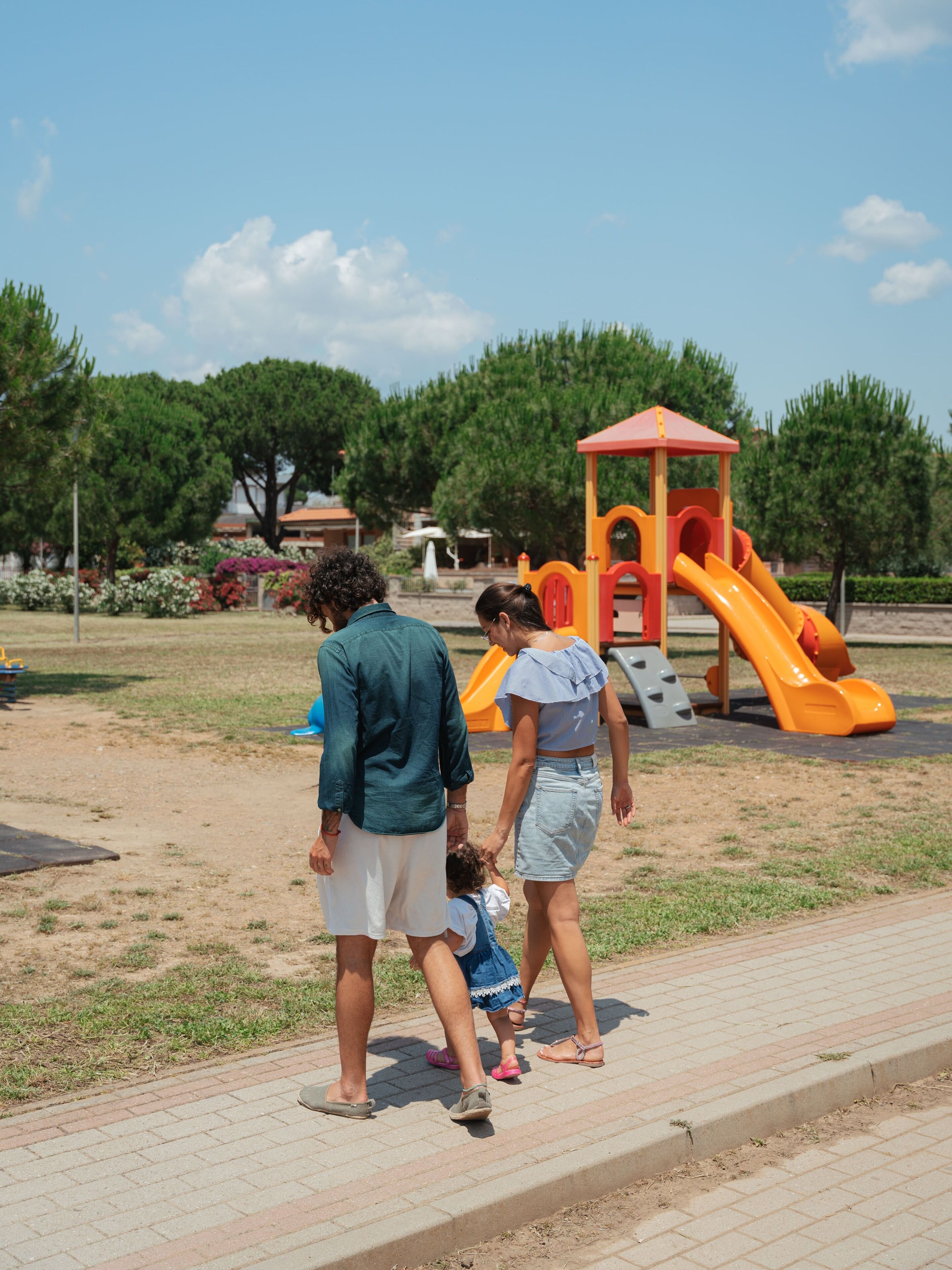 A playground with a slide and climbing structures, surrounded by trees and a paved walkway.