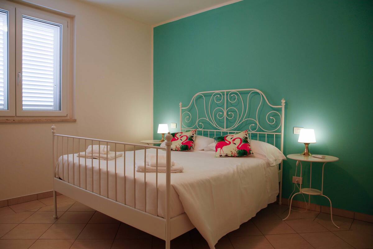 Un lit bien fait, avec une literie blanche et des coussins décoratifs, une tête de lit en métal, une table de chevet avec une lampe et une fenêtre équipée de stores.