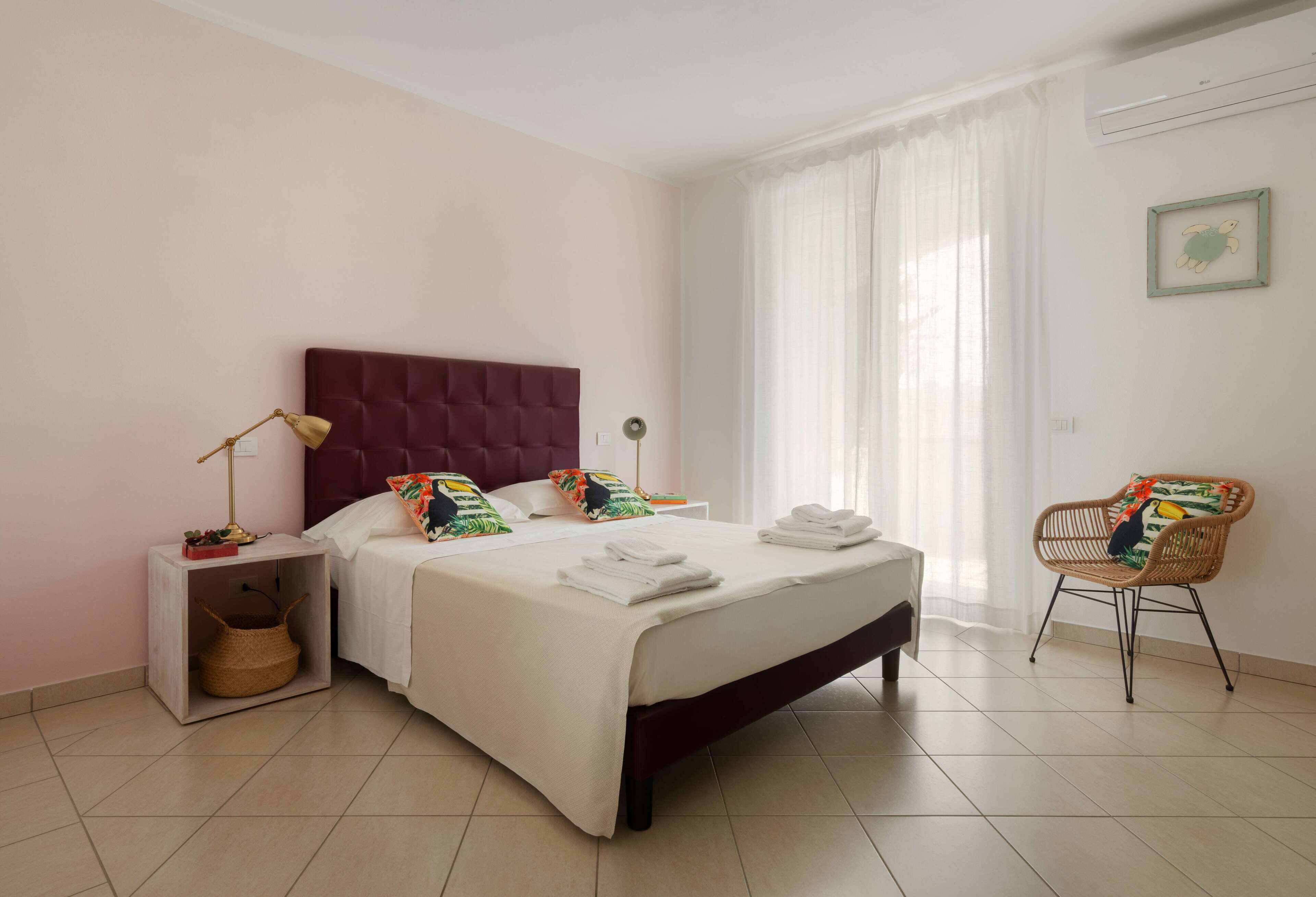 A neatly made bed with white linens, a red headboard, and colorful pillows. There is a wicker chair with a cushion and a small table with a lamp and a basket.