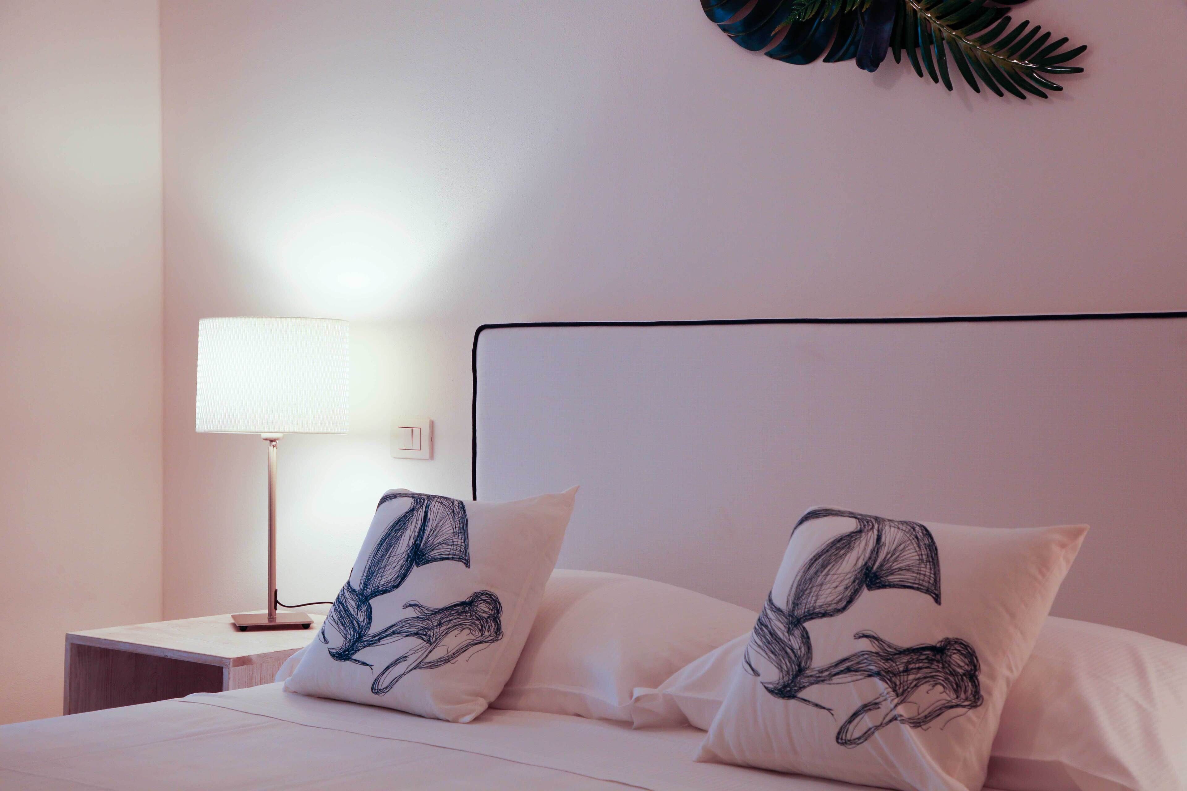 Un lit bien fait, avec deux oreillers ornés d’un motif de feuilles, une lampe de chevet et une plante fixée au mur.