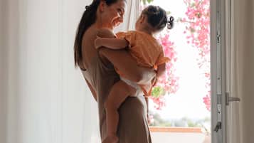 A person holding a child in a room with a large window and a view of pink flowers outside.
