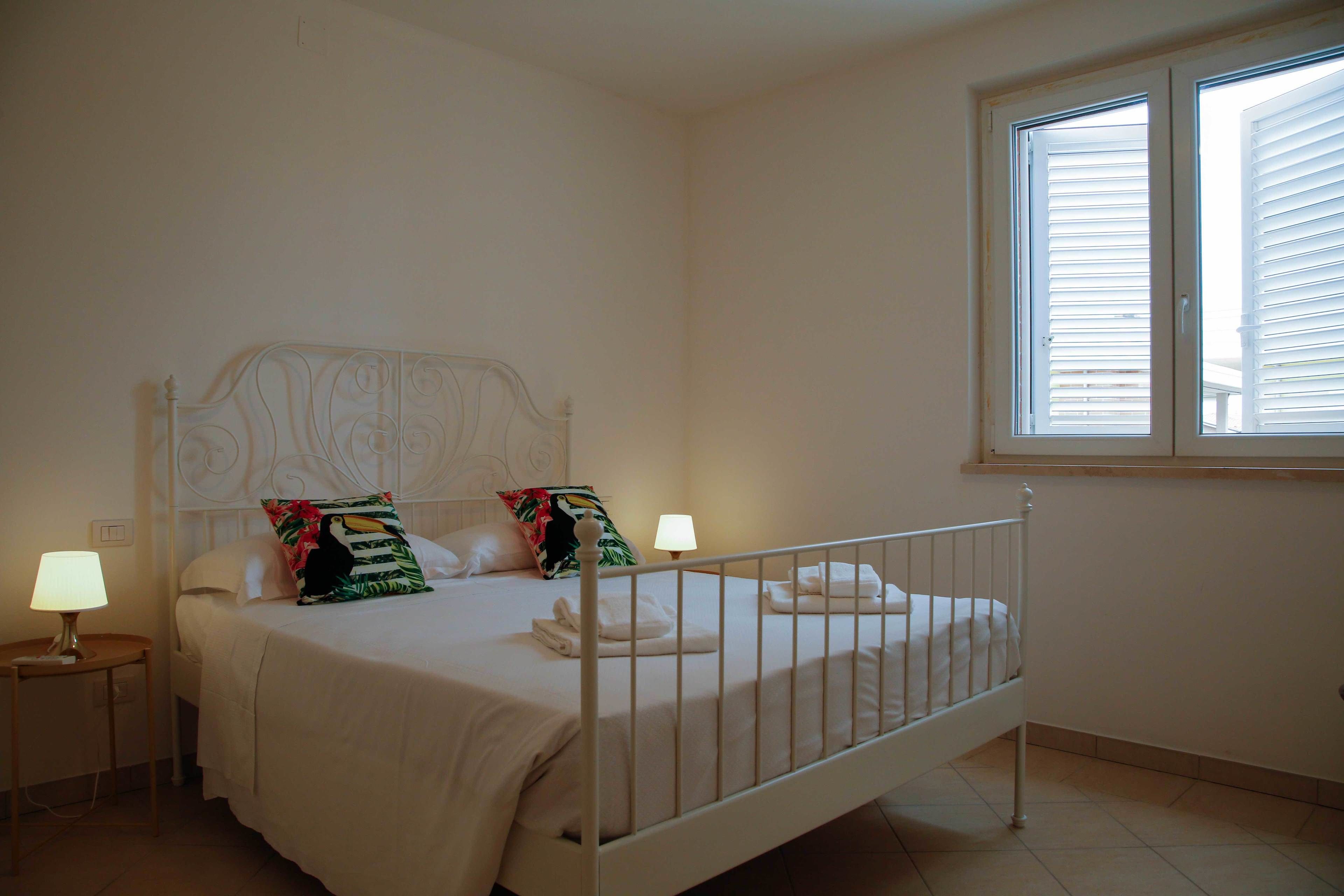 A neatly made bed with white linens and two pillows, a bedside lamp, and a window with blinds.