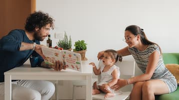 Eine Familie liest gemeinsam ein Buch an einem weißen Tisch in einem Raum mit einem grünen Sofa und einer kleinen Pflanze auf dem Tisch.