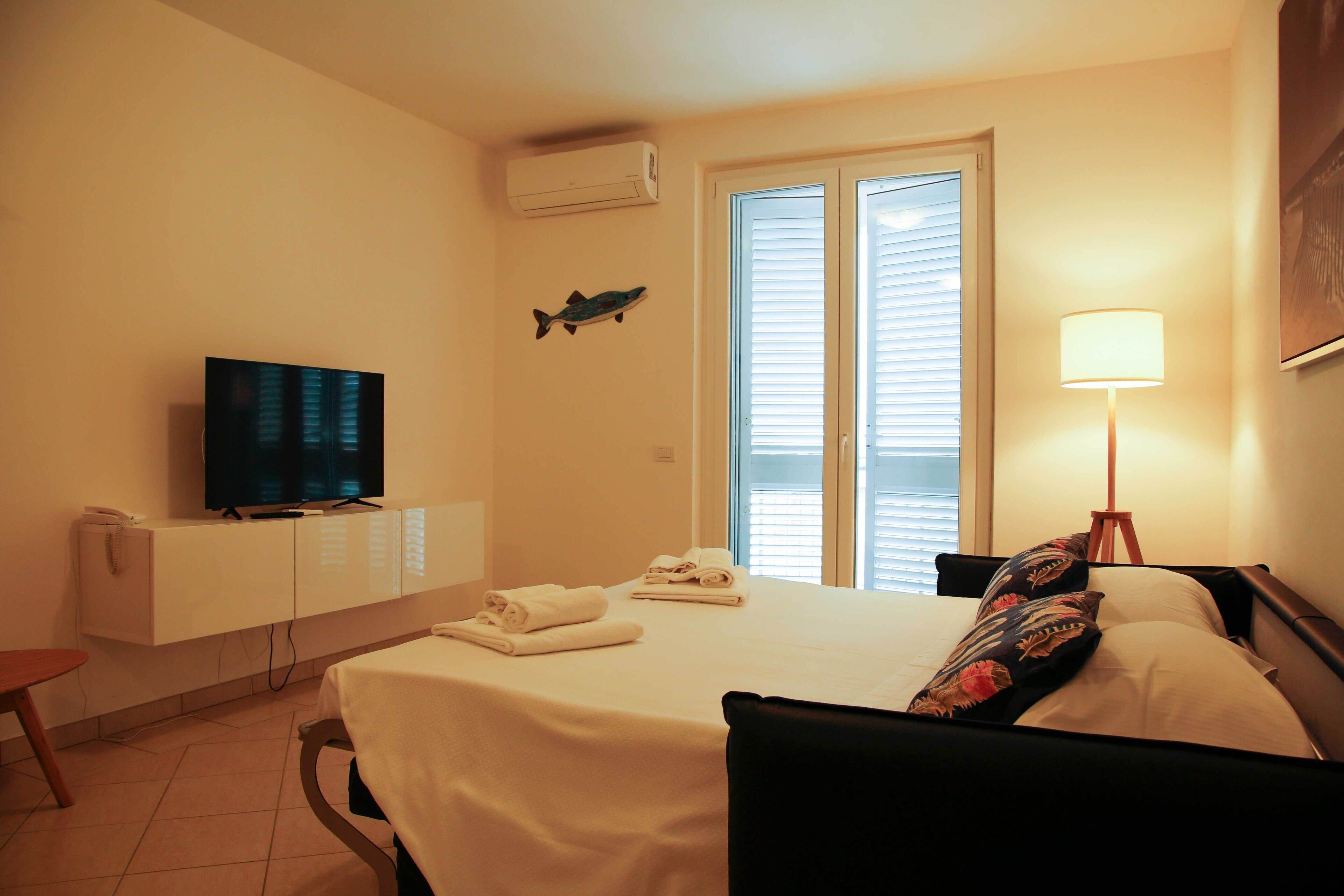 A hotel room with a bed, a TV, a desk, a lamp, and a window with blinds.