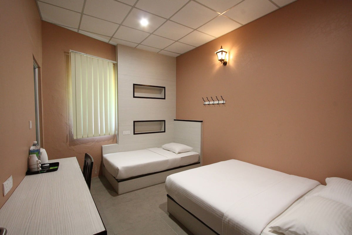 A hotel room with two single beds, a small table, a chair, a window with blinds, a wall-mounted light, and a coat rack.