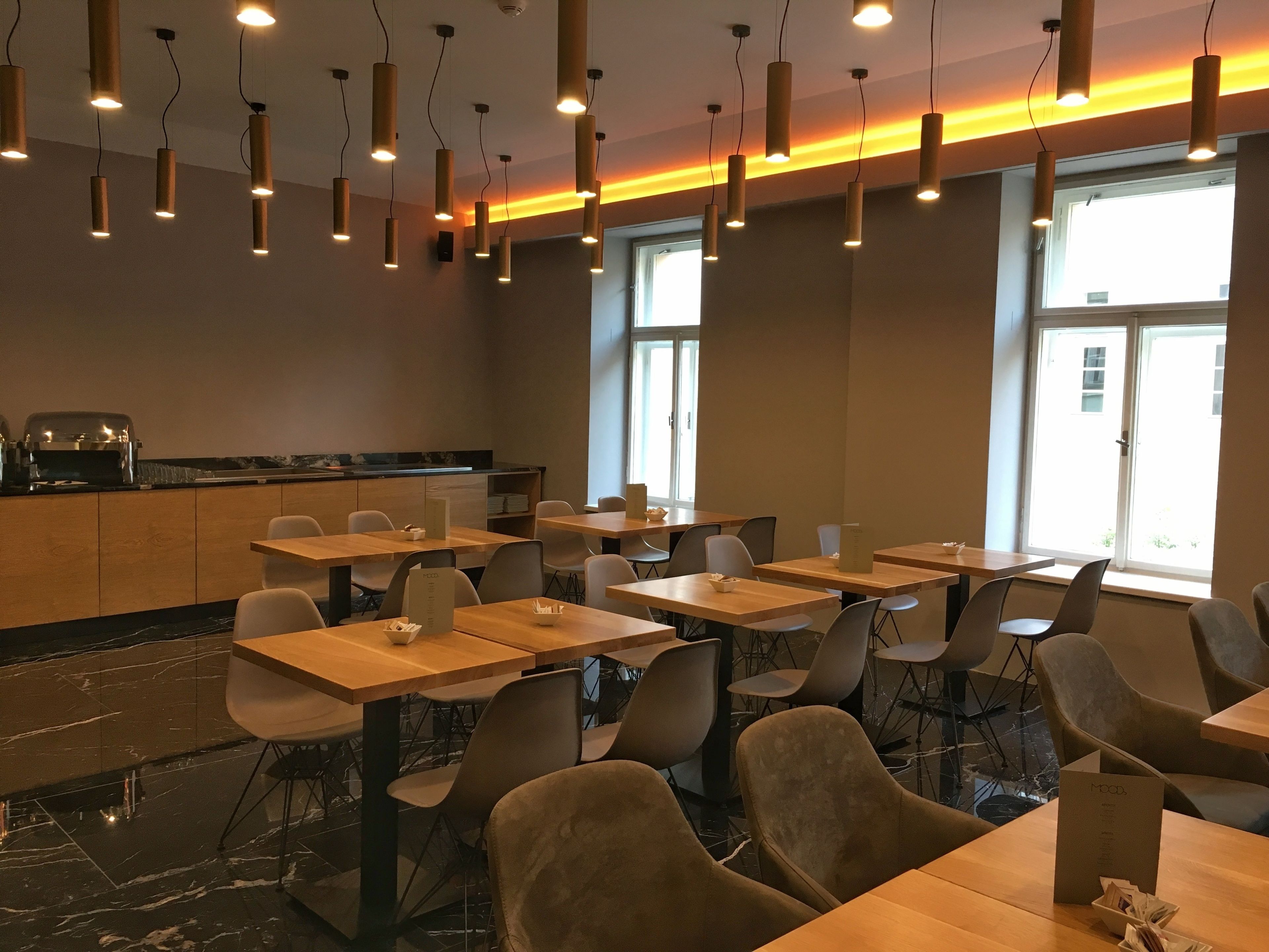 Interior moderno de restaurante com várias mesas e cadeiras, iluminação pendente, balcão com caixa registradora e grandes janelas que proporcionam luz natural.