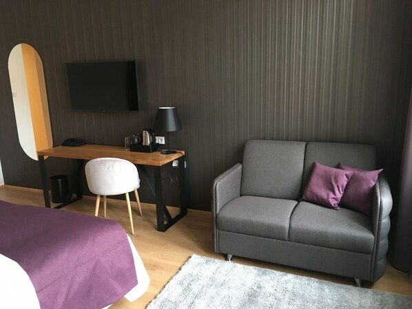 Una habitación moderna con un escritorio de madera, un sofá gris con un cojín morado, una silla blanca y un televisor de pantalla plana montado en la pared.