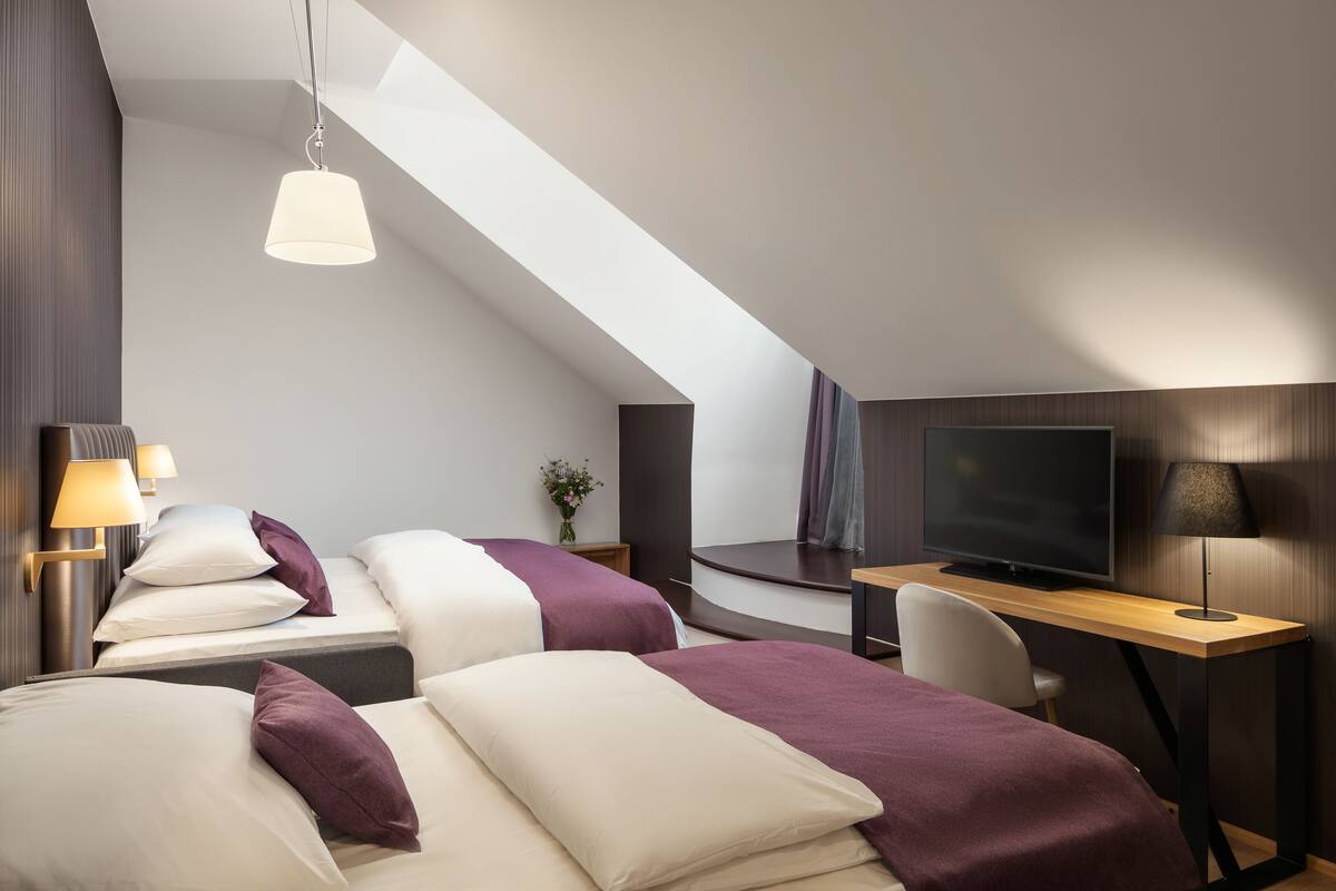 A modern hotel room with two beds, a desk, a TV, and a skylight.