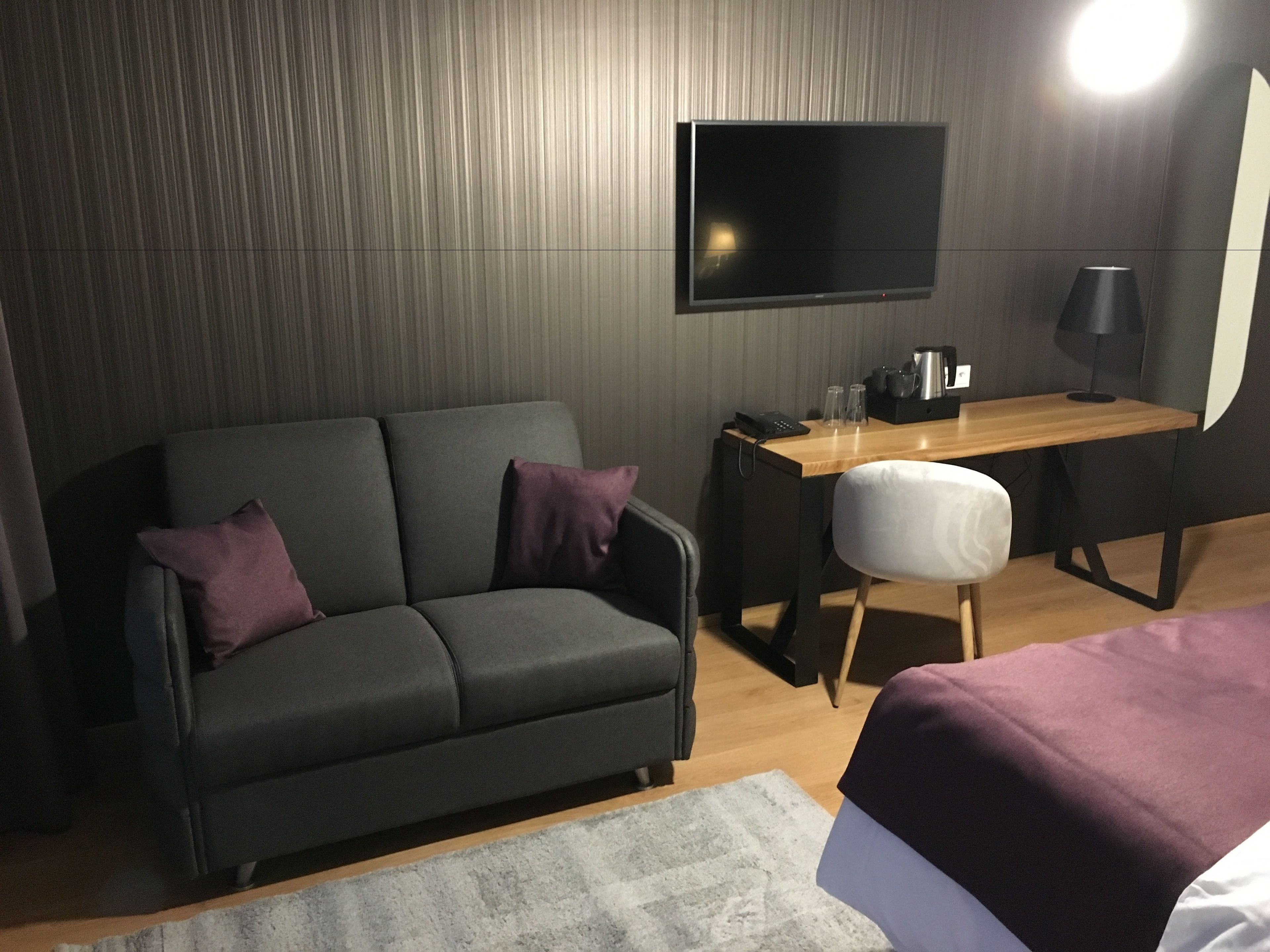 Een moderne hotelkamer met een donkergrijze bank, een klein houten bureau, een flatscreen televisie aan de wand en een bed met een paars sprei.