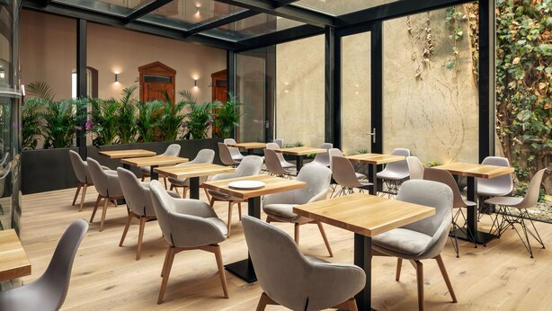 Un espace de restauration intérieur moderne, avec des tables en bois et des chaises à revêtement gris, de grandes fenêtres et des plantes d’intérieur.