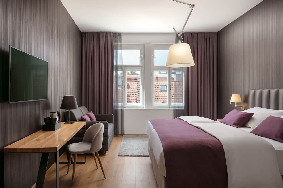A modern hotel room with a bed, a desk, a chair, a TV, and a window with curtains.