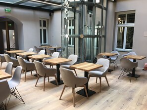 Un espace de restauration moderne avec des tables en bois et des chaises grises, de grandes fenêtres et un puits d’ascenseur en verre.