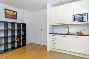 Una cocina moderna con armarios blancos, un microondas y un lavabo. Se observa una estantería negra con objetos decorativos y una puerta que da a otra habitación.