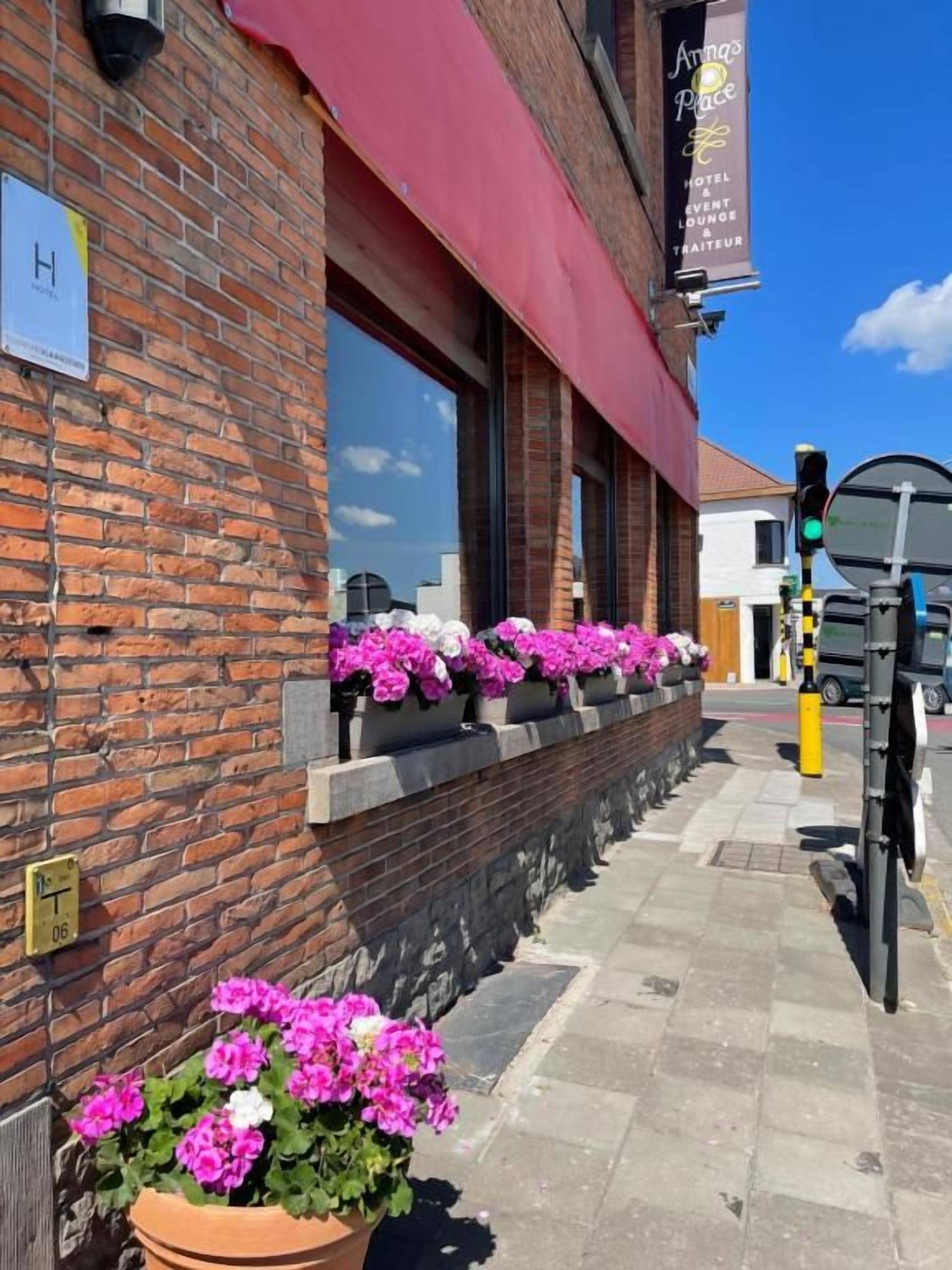 Een gebouw van baksteen met een rode luifel, bloembakken met roze bloemen en een helderblauwe lucht.
