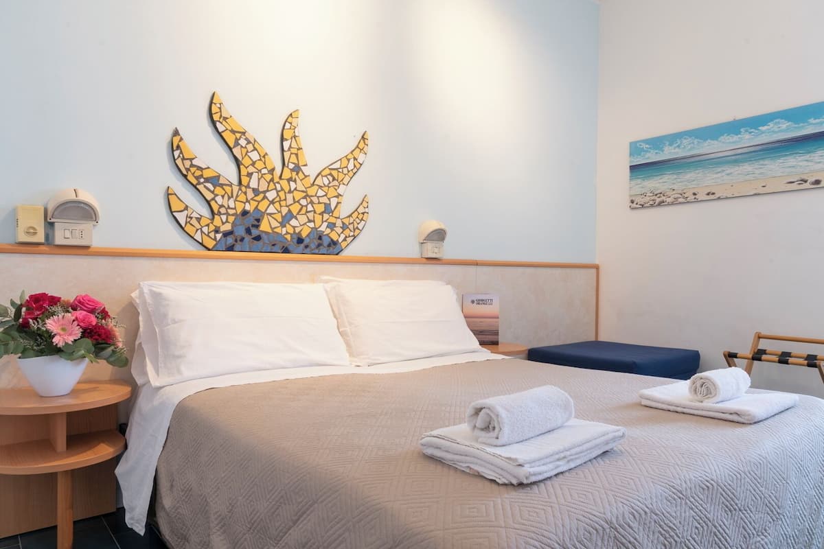 Una camera d'albergo con un letto, un comodino, una lampada a parete, un quadro e un vaso di fiori.
