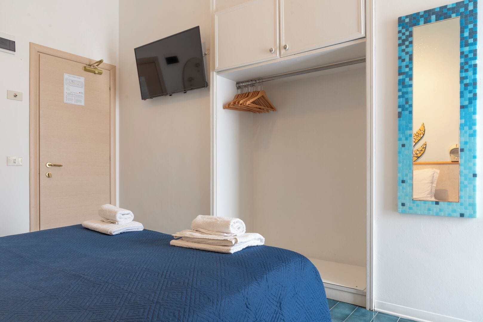 Une chambre d’hôtel avec un lit, un téléviseur fixé au mur, un miroir avec un cadre en mosaïque et une porte dotée d’un porte-serviettes.