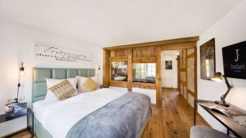 A modern bedroom with a large bed, a wooden wardrobe, a desk with a lamp, and a wall-mounted artwork.