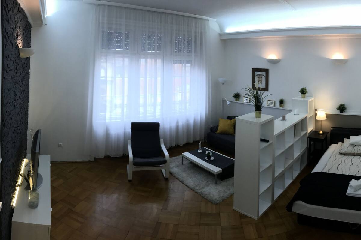 Une chambre d’hôtel moderne équipée d’un lit, de tables de chevet, d’un bureau, d’une chaise, d’une fenêtre avec des rideaux légers et d’une petite plante.