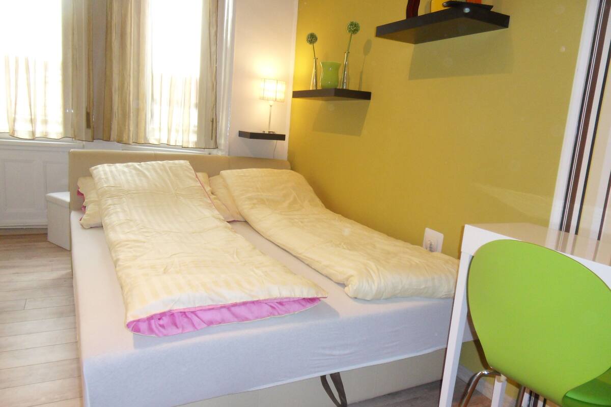 Un lit simple avec un matelas blanc et une couverture rose, une chaise verte et une table de chevet avec une lampe.