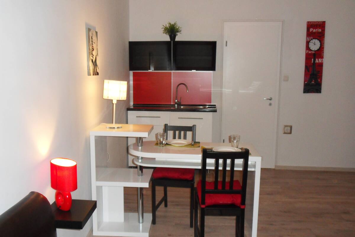 Une cuisine compacte dotée d’un plan de travail blanc, de placards rouges et noirs, d’un évier et d’un coin repas avec une table et des chaises blanches.