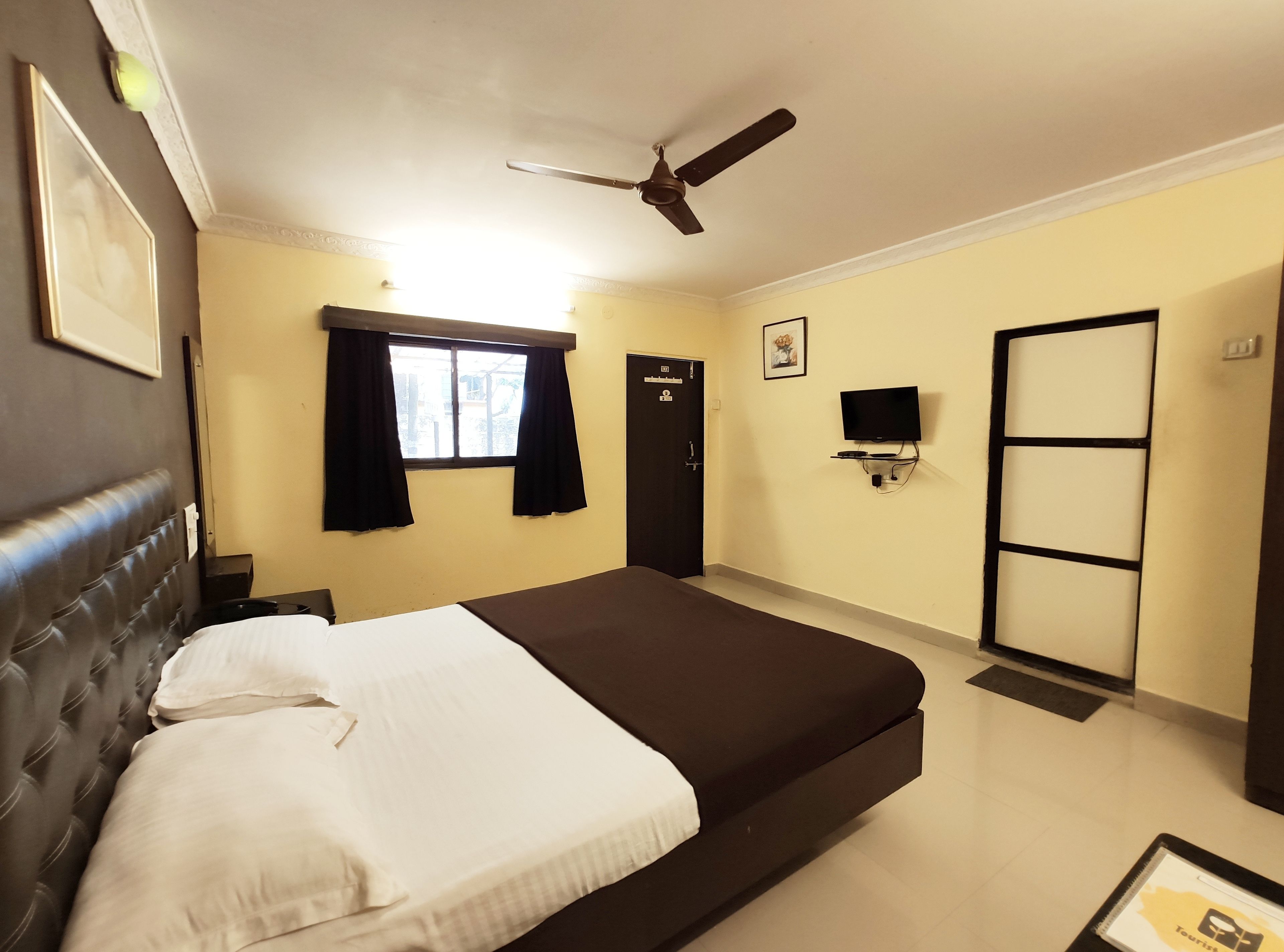 Una habitación de hotel con cama, ventilador de techo, televisor montado en la pared y ventana con cortinas.