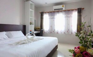 A neatly made bed with white linens and a heart-shaped towel arrangement, a bedside table with a vase of flowers, and a window with sheer curtains.