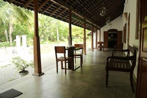 A covered outdoor area with a table and chairs, surrounded by greenery.