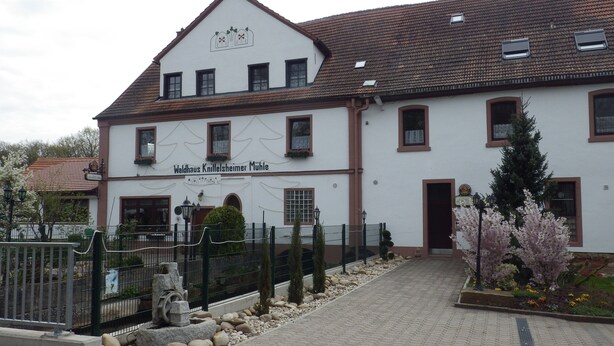 Un edificio tradicional con un letrero que dice "Waldhaus Knittelheimer Mühle" y un área de acceso pavimentada con una cerca y plantas decorativas.