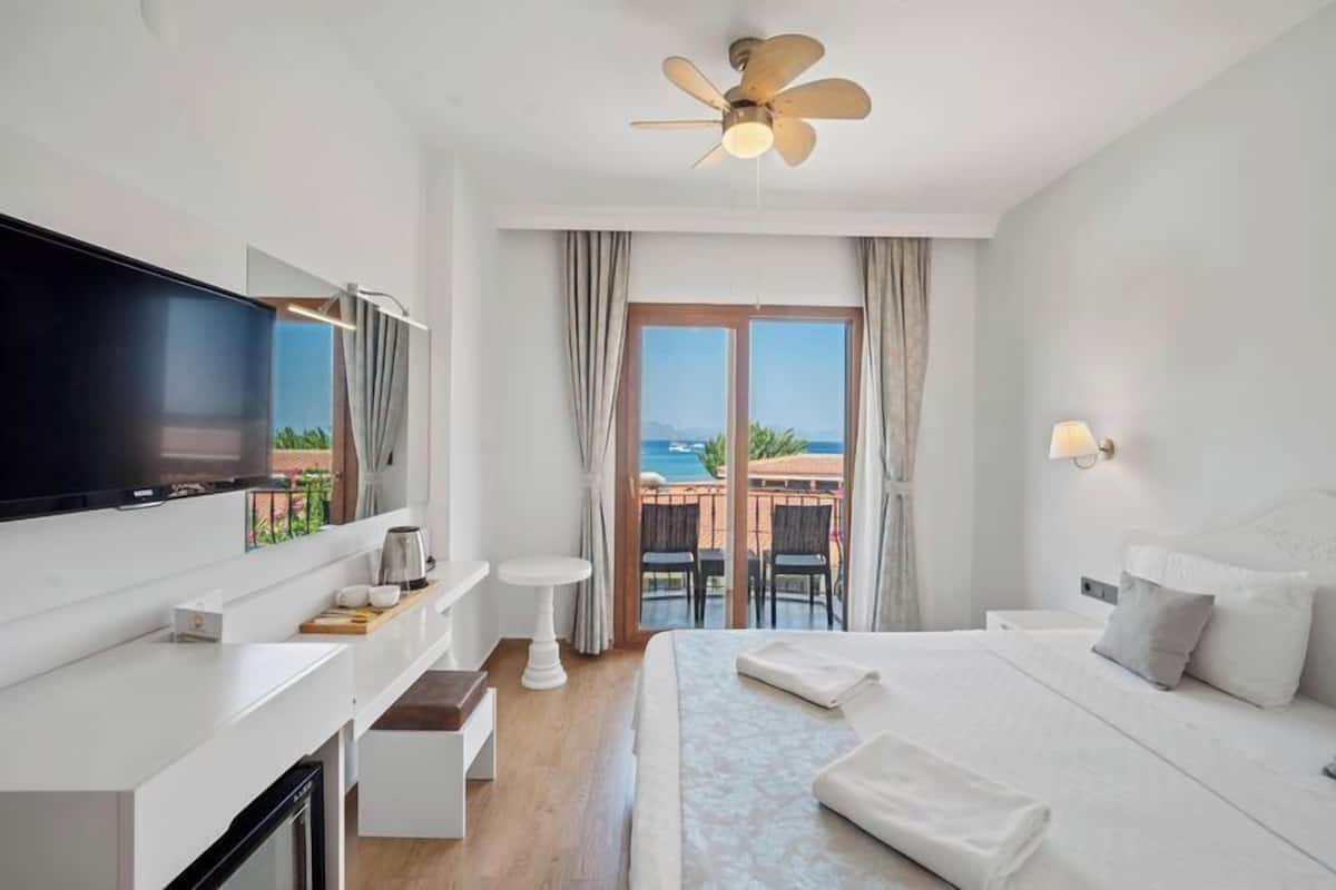 Habitación de hotel con una cama grande, un televisor de pantalla plana, un ventilador de techo, un balcón con vista al mar y una pequeña mesa con cafetera.