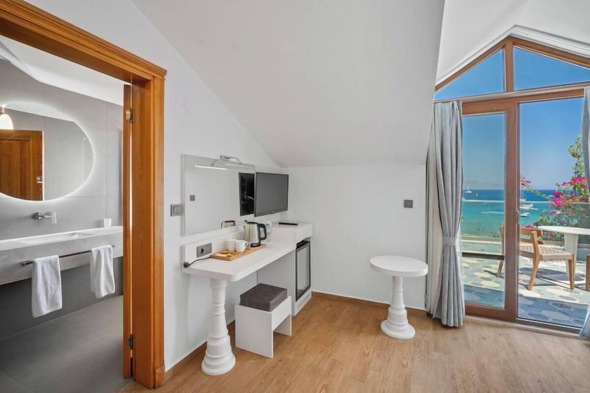 Un baño moderno con un espejo grande, un lavabo y un inodoro. Hay una mesita con una tetera y tazas. La habitación tiene suelo de madera y un ventanal con vistas al océano.