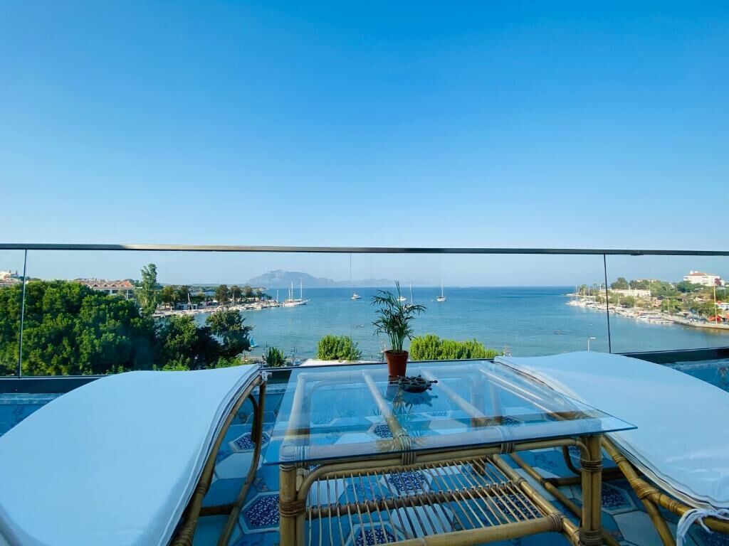 Balcón con mesa de vidrio y silla de mimbre con vista a una zona costera con barcos y vegetación.
