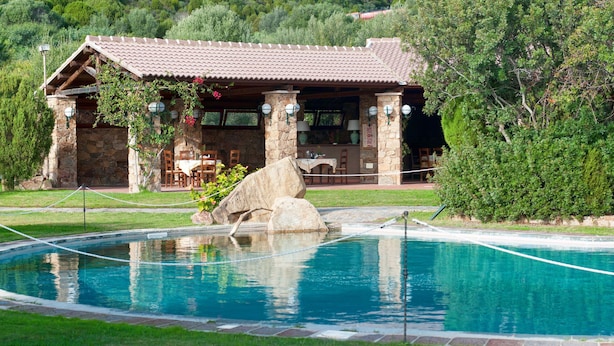 A stone building with a tiled roof, a pool with a rope barrier, and a stone wall with a rock feature. - Hotel Spartivento (Domus de Maria)