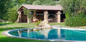 A stone building with a tiled roof, a pool with a rope barrier, and a stone wall with a rock feature. - Hotel Spartivento (Domus de Maria)