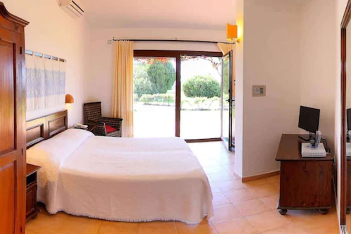 Ein Hotelzimmer mit einem großen Bett, einem Schreibtisch mit Computer, einem Stuhl und Blick ins Freie durch eine Schiebetür.