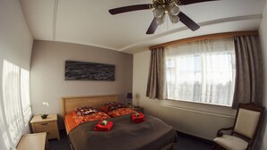 Une chambre à coucher comprenant un lit, une chaise, une table de chevet, une fenêtre avec des rideaux, un ventilateur de plafond et un radiateur.