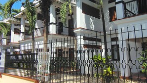 A multi-story building with balconies, surrounded by a black wrought iron fence, and palm trees in the foreground. - Mangro Hotel Diani Beach (Ukunda)