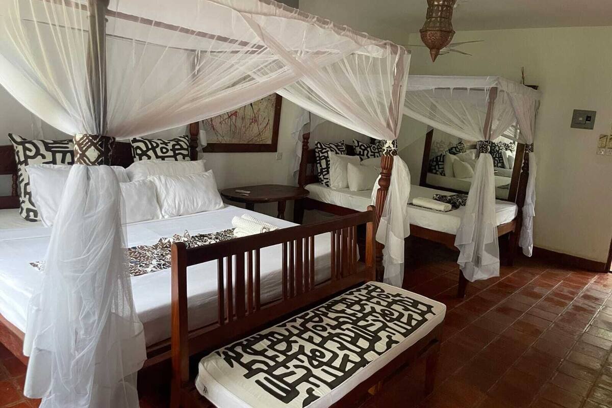 A room with two beds, each with a canopy. There is a bench in front of the beds, and a small table beside the left bed.